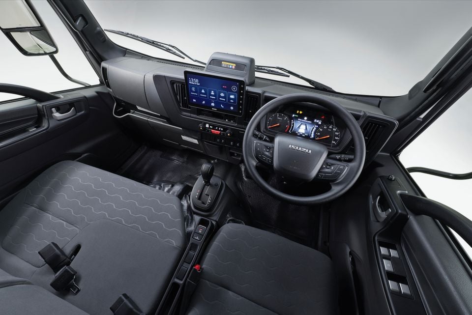 The cabin of a 2026 Isuzu NMS truck, showing the steering wheel, digital instrument cluster, touchscreen infotainment display and front seats