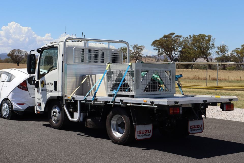A white Isuzu flatbed truck fitted for ANCAP crash testing, seen from the rear quarter with test equipment strapped to its tray.