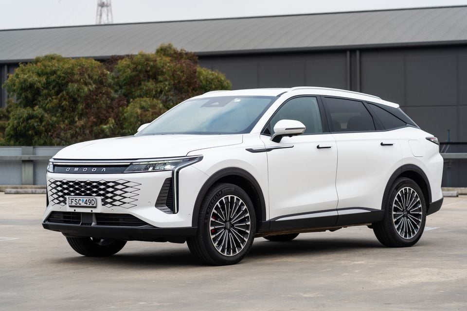 A white Omoda 9 SUV seen from the front-left side, parked in an open area with a building and trees in the background.