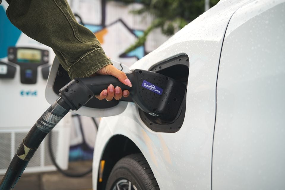 A person plugs a charging connector into the charging port of a white 2026 MG4 EV electric hatchback in the rain.