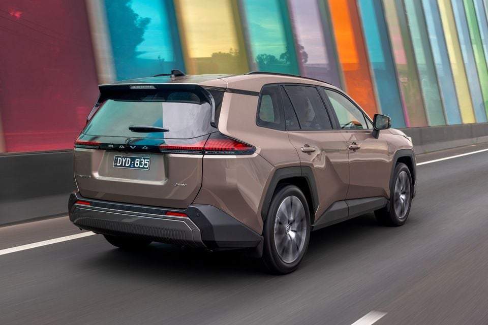 A bronze Toyota RAV4 Cruiser hybrid SUV seen from the rear three-quarter angle while driving on a road, with a colourful striped wall in the background