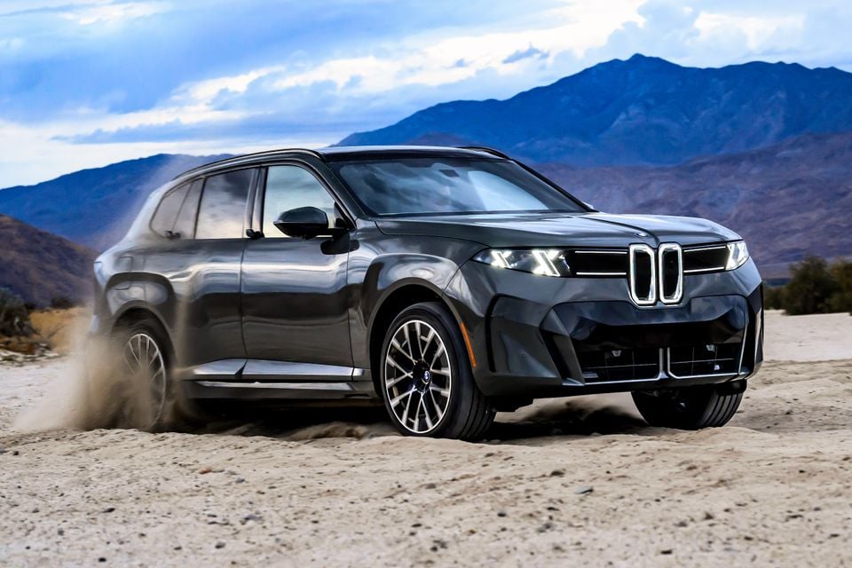 A dark grey 2027 BMW iX5 60 xDrive SUV kicking up sand as it drives across a desert with mountains behind