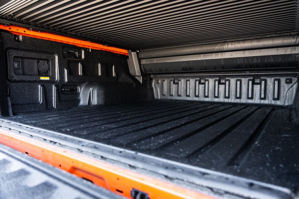 The lined tub of a 2027 Ford Ranger Wildtrak ute, showing the bed liner, tie-down points, power outlets on the front wall, and a roller cover, with orange accents on the body sides