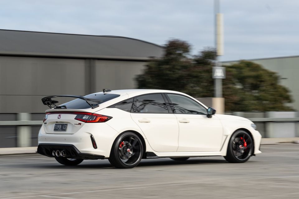 A white Honda Civic Type R hatchback seen from the rear three-quarter angle while driving in a car park, showing its large rear wing, triple exhaust outlets, and red brake callipers