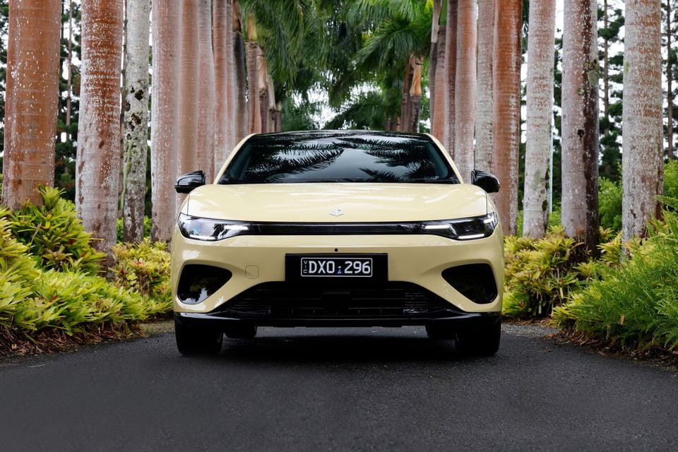 A pale yellow 2027 Leapmotor B05 hatchback seen from the front, parked on a road lined with palm trees
