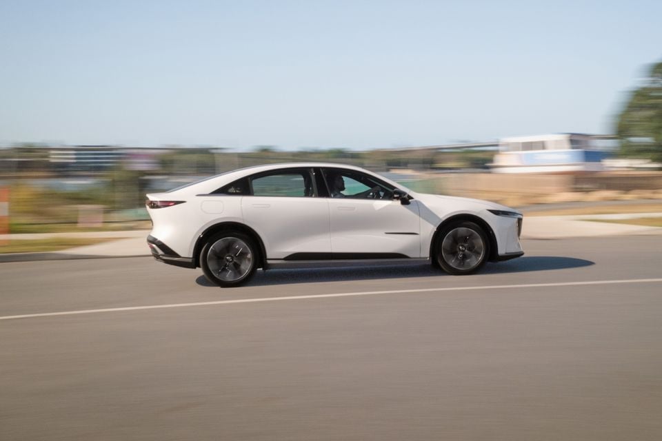 A white 2027 Mazda 6e Atenza sedan driving on a road, seen from the side with motion blur in the background