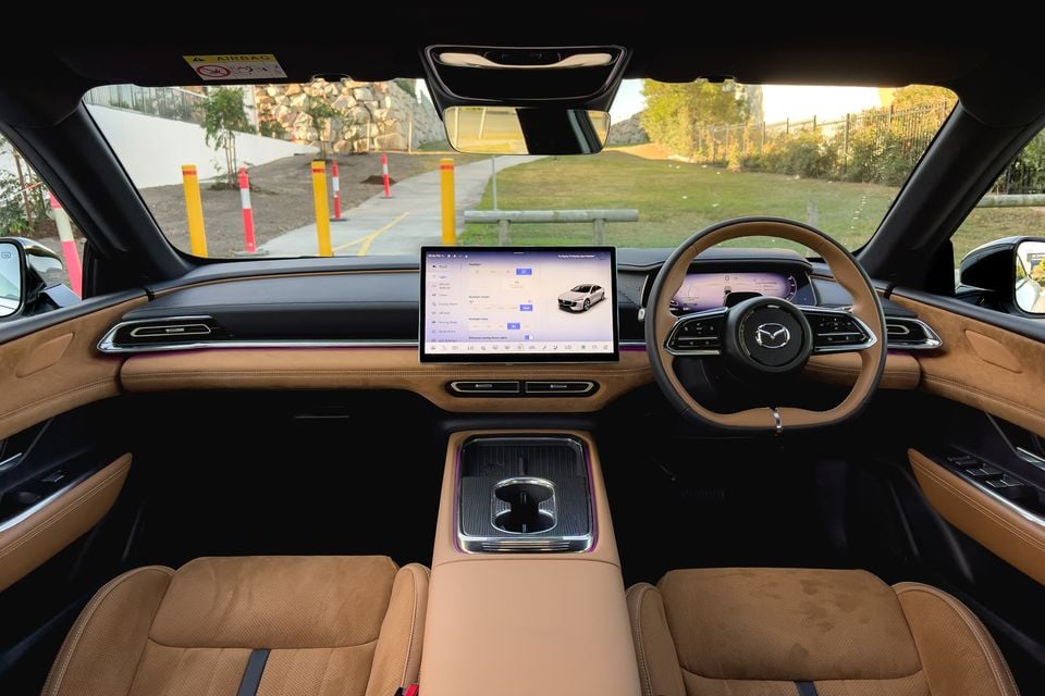 The tan leather interior of a 2027 Mazda 6e Atenza, viewed from the rear seats showing the dashboard, steering wheel and centre touchscreen