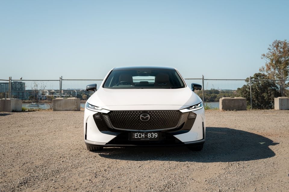 A white 2027 Mazda 6e Atenza sedan parked facing the camera near a waterfront, with its front grille and headlights clearly visible.