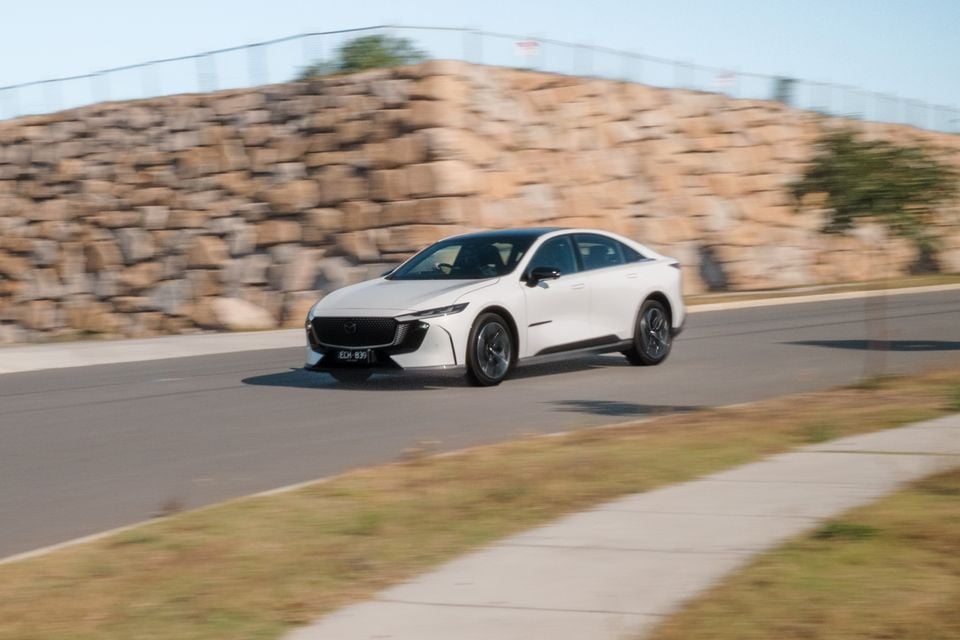 A white 2027 Mazda 6e Atenza sedan driving on a road, viewed from the front quarter with motion blur in the background.