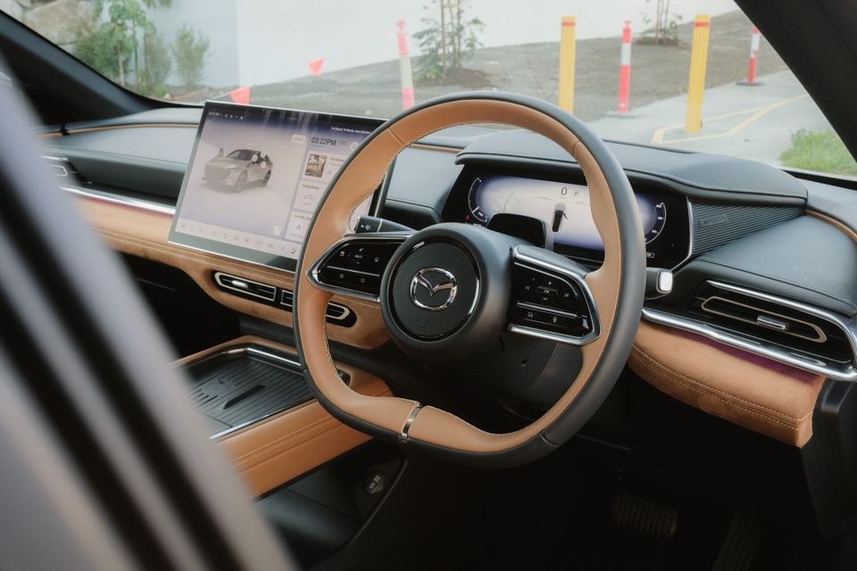 The steering wheel, digital instrument cluster and infotainment screen inside a 2027 Mazda 6e Atenza, with a parking area visible through the windscreen.