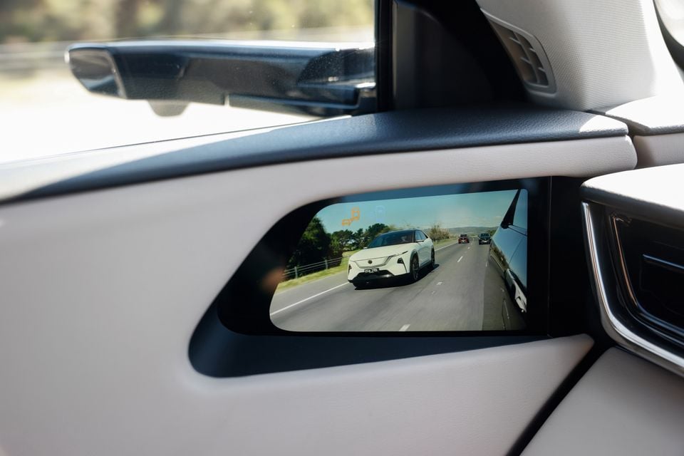A digital side mirror screen inside a Mazda CX-6e shows a camera view of a following white SUV on a highway.