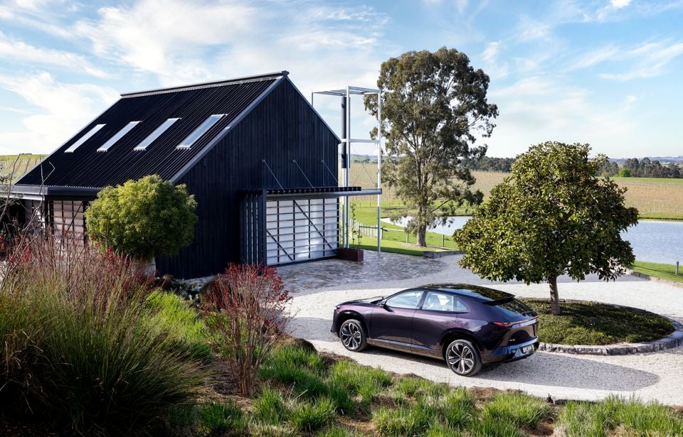 A purple 2027 Mazda CX-6e SUV parked on a gravel driveway beside a modern black barn-style house with a lake in the background.