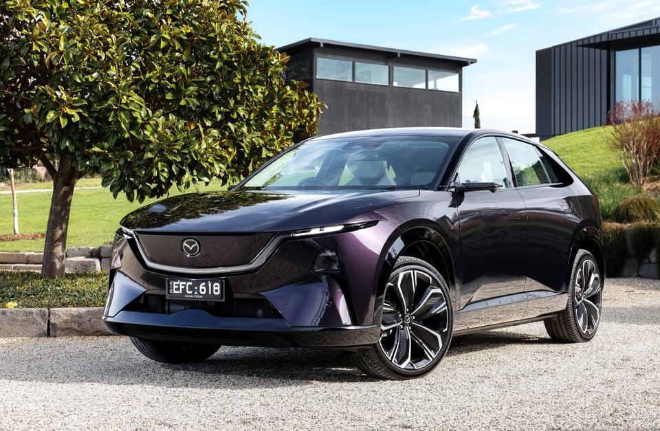 A dark purple 2027 Mazda CX-6e SUV parked on gravel in front of a modern house, seen from the front quarter.