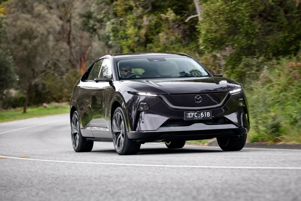 A dark purple 2027 Mazda CX-6e electric SUV driving on a country road, seen from the front quarter angle