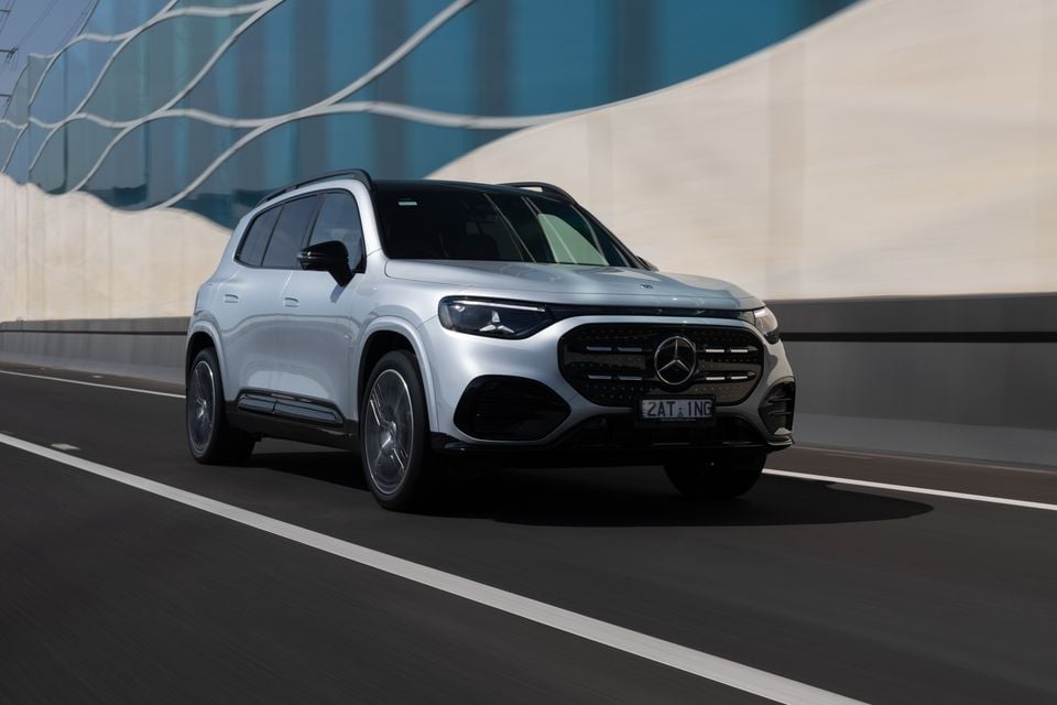 A silver 2027 Mercedes-Benz GLB 350 4Matic electric SUV driving along a road past a curved concrete wall, seen from the front.