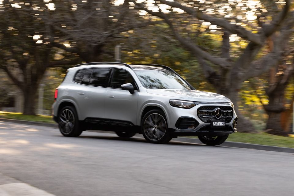 A silver 2027 Mercedes-Benz GLB 350 4Matic electric SUV driving along a tree-lined street, seen from the front three-quarter angle