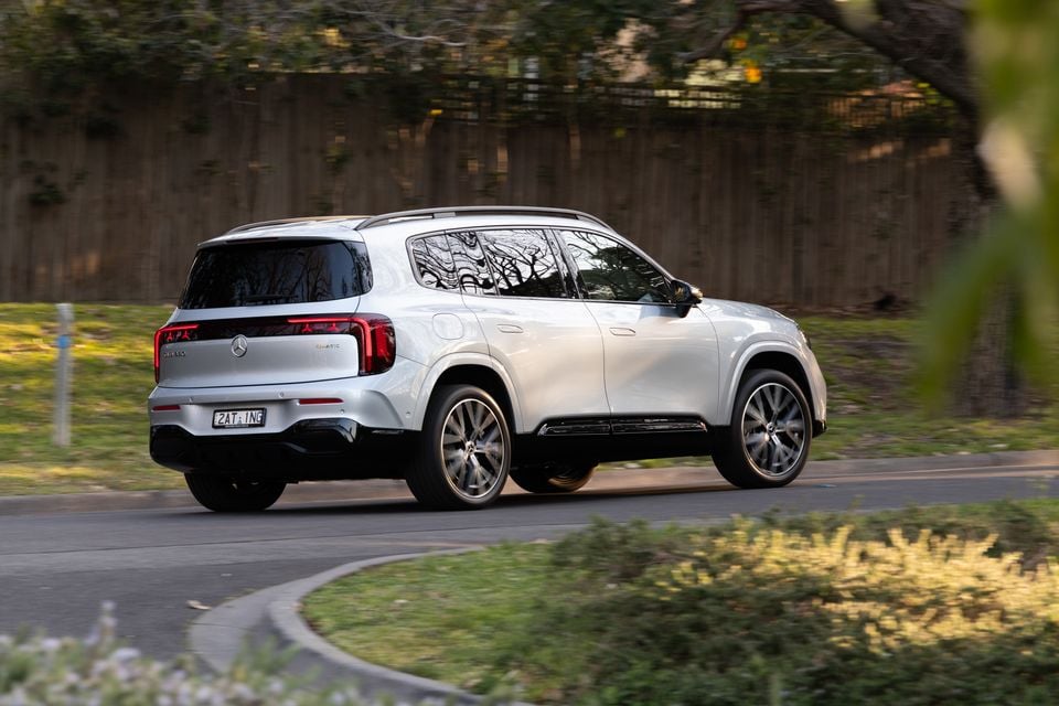 A silver 2027 Mercedes-Benz GLB 350 4Matic electric SUV driving along a suburban road, seen from the rear passenger side