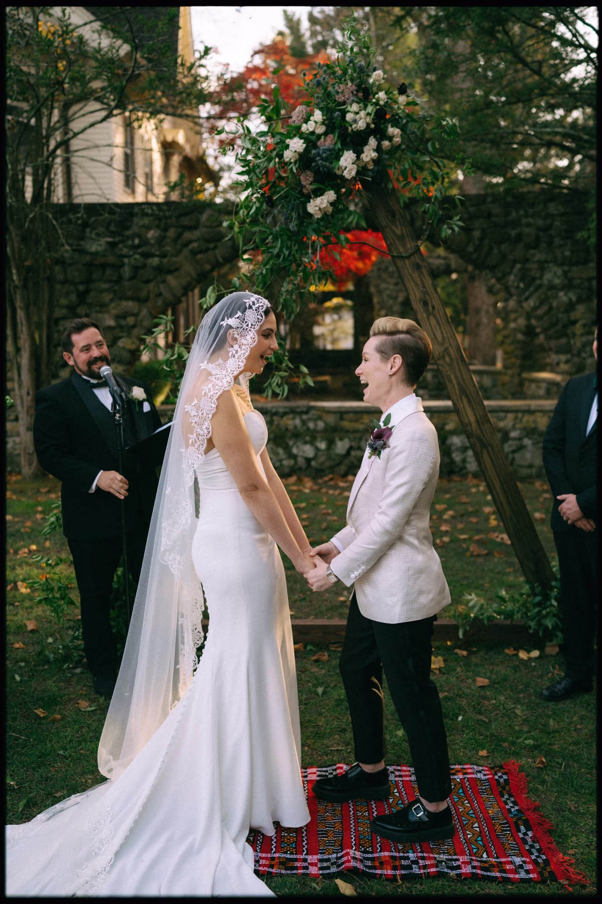 LGBTQ Couple holding hands and laughing beneath a triangular wooden ceremony arch decorated with flowers and foliage