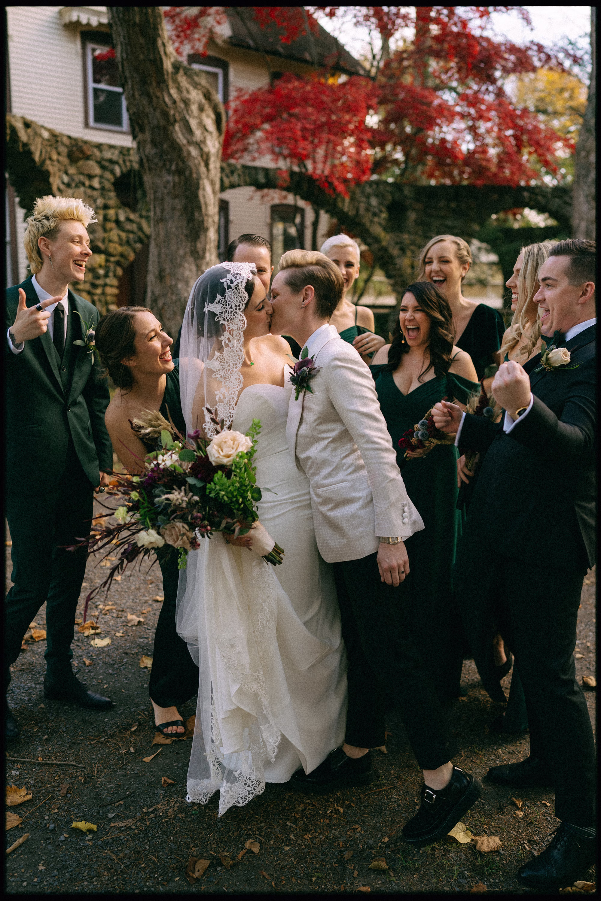LGBTQ Newlyweds kissing amid their cheering wedding party beneath red autumn leaves