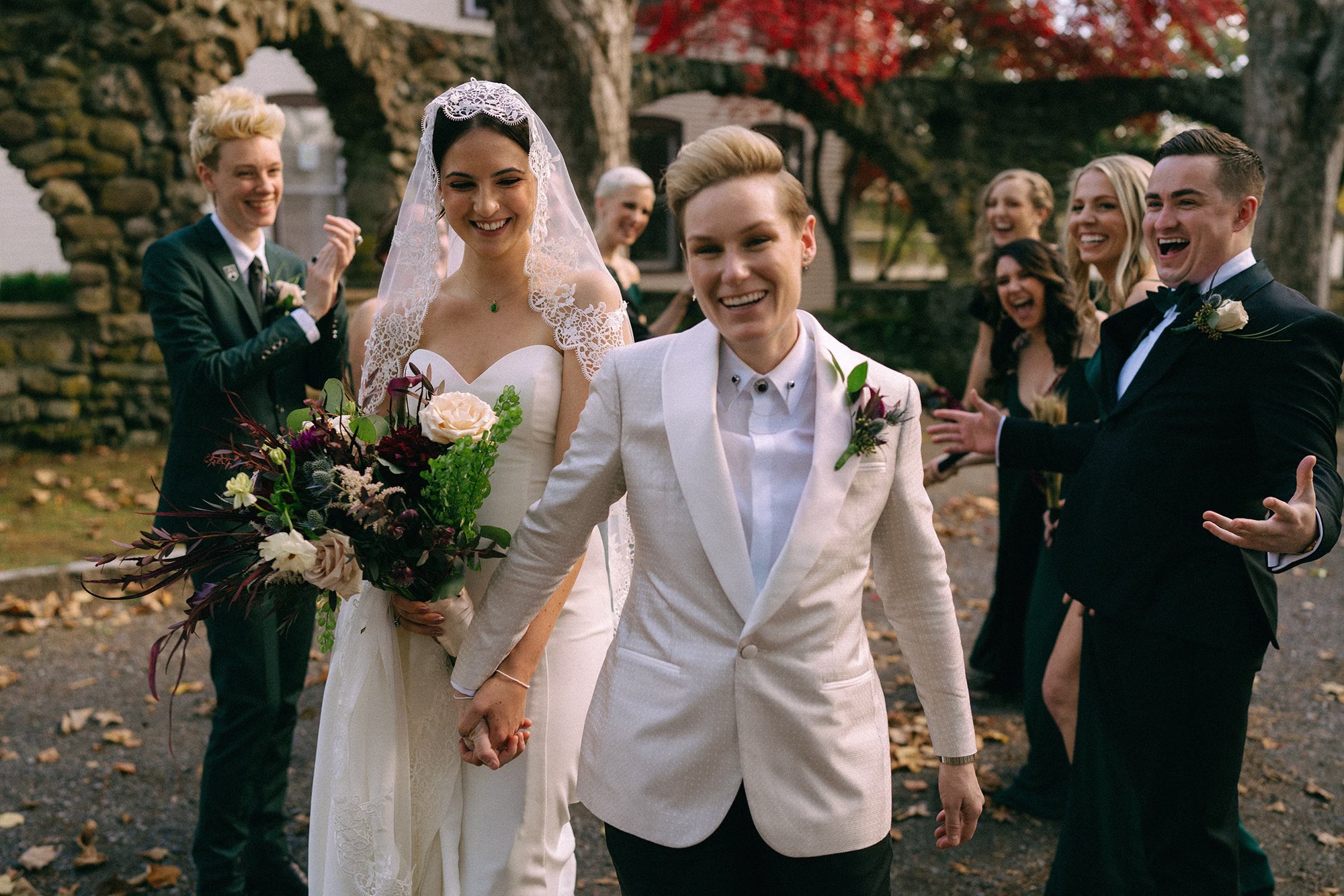 LGBTQ Newlyweds holding hands outside stone arches as their wedding party cheers around them