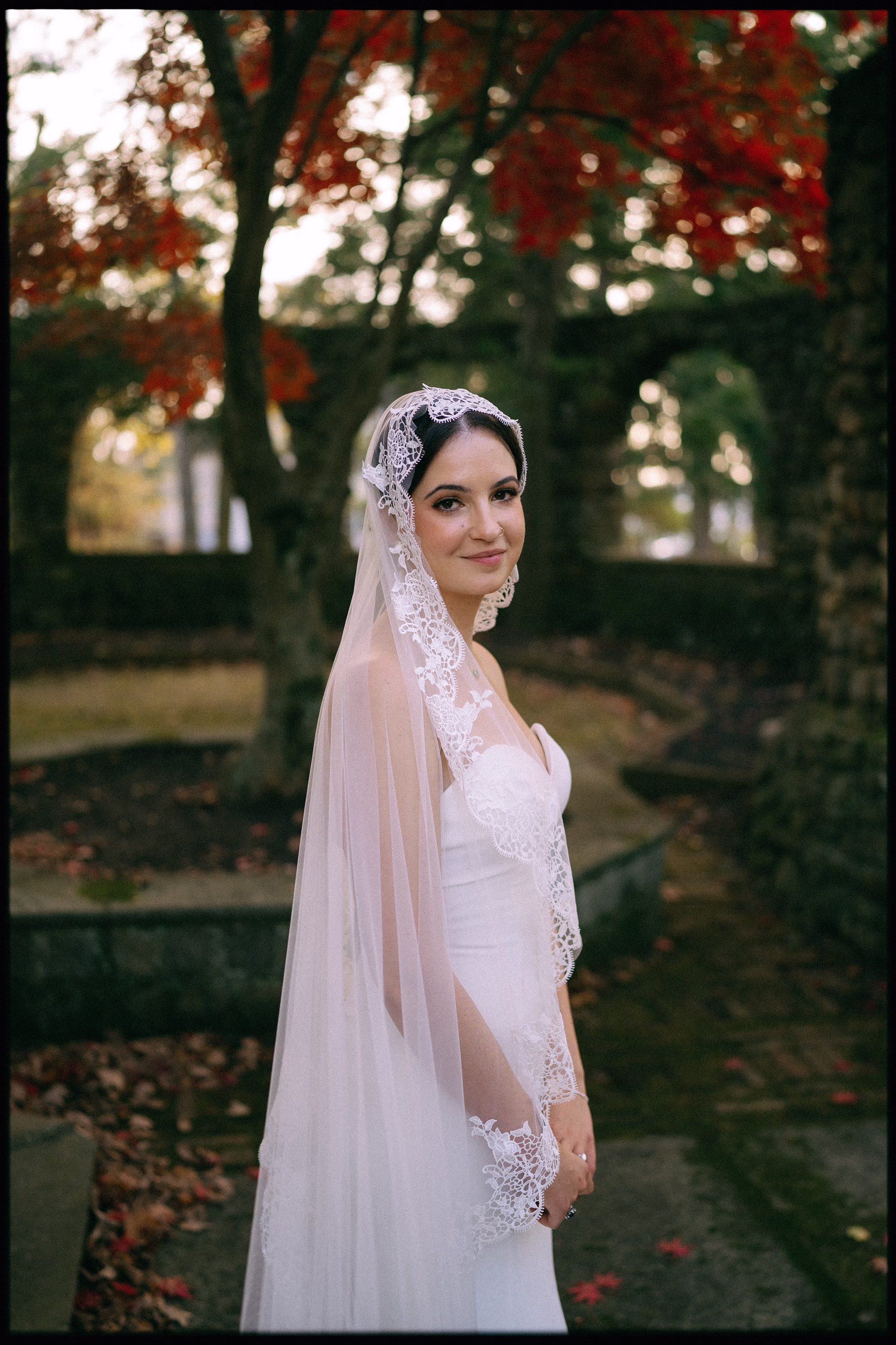 Bride wearing the strapless Leona gown by Casablanca Bridal and a long floral lace-edged veil beneath autumn trees