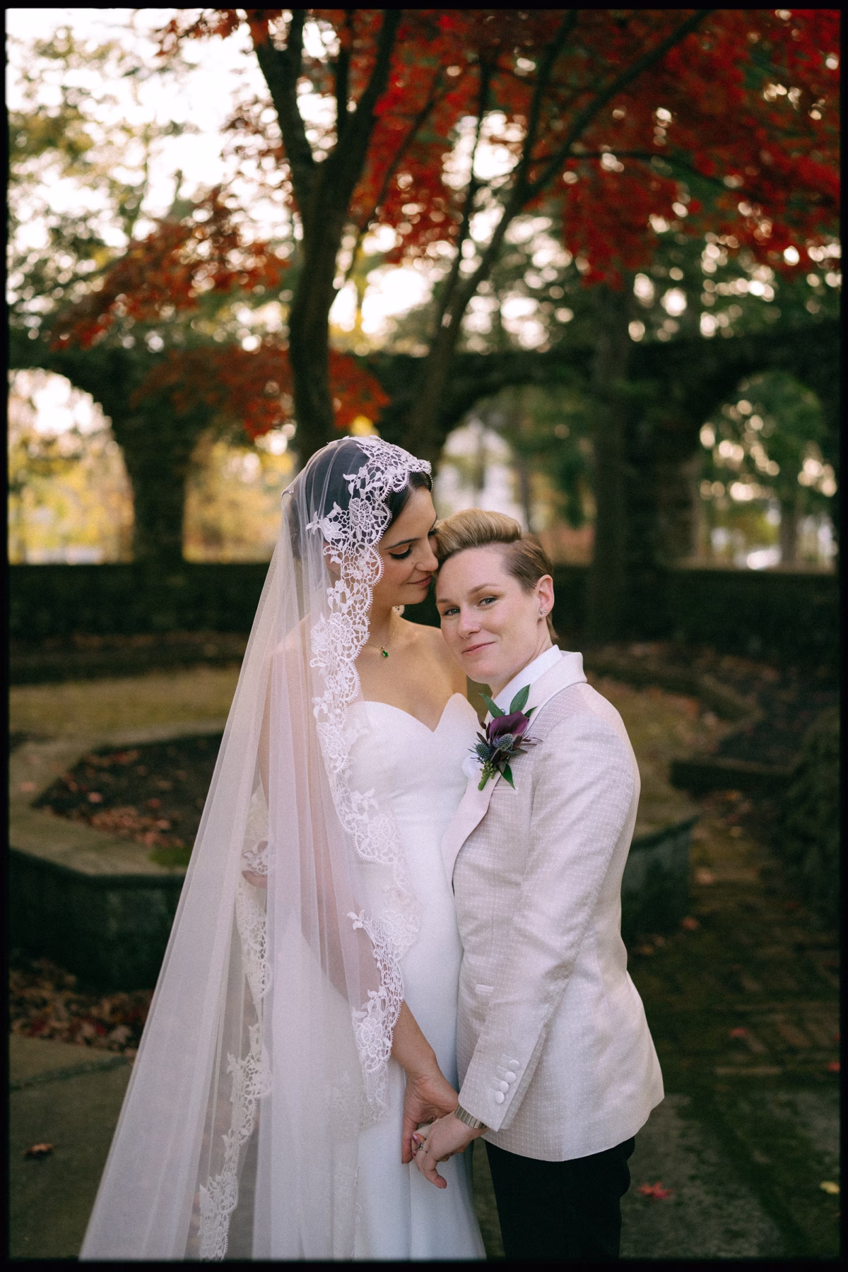 Couple embracing beneath autumn leaves, one wearing a Casablanca Bridal gown and lace-edged veil and the other a white tuxedo jacket
