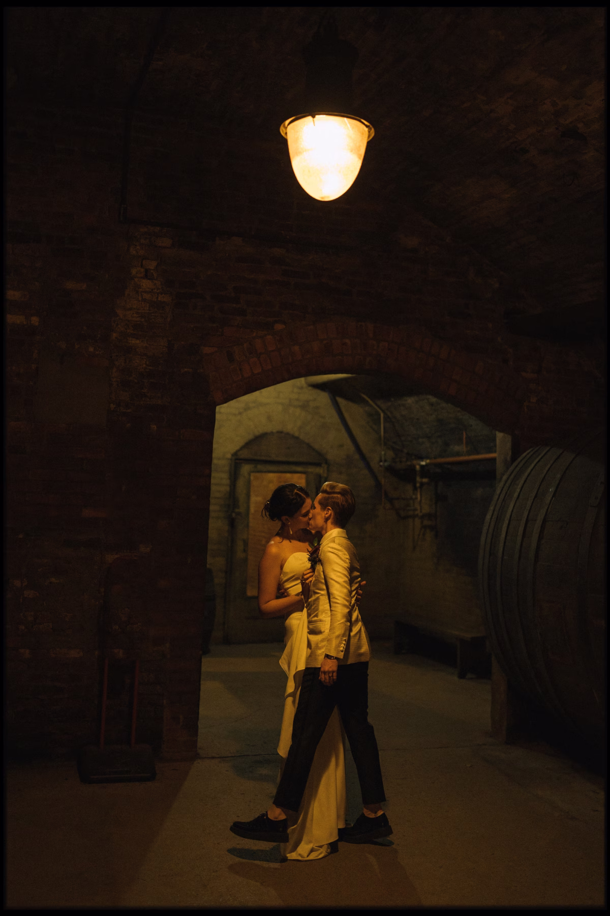 Couple kissing beneath a brick arch beside large wooden barrels in Brotherhood Winery’s dimly lit cellar