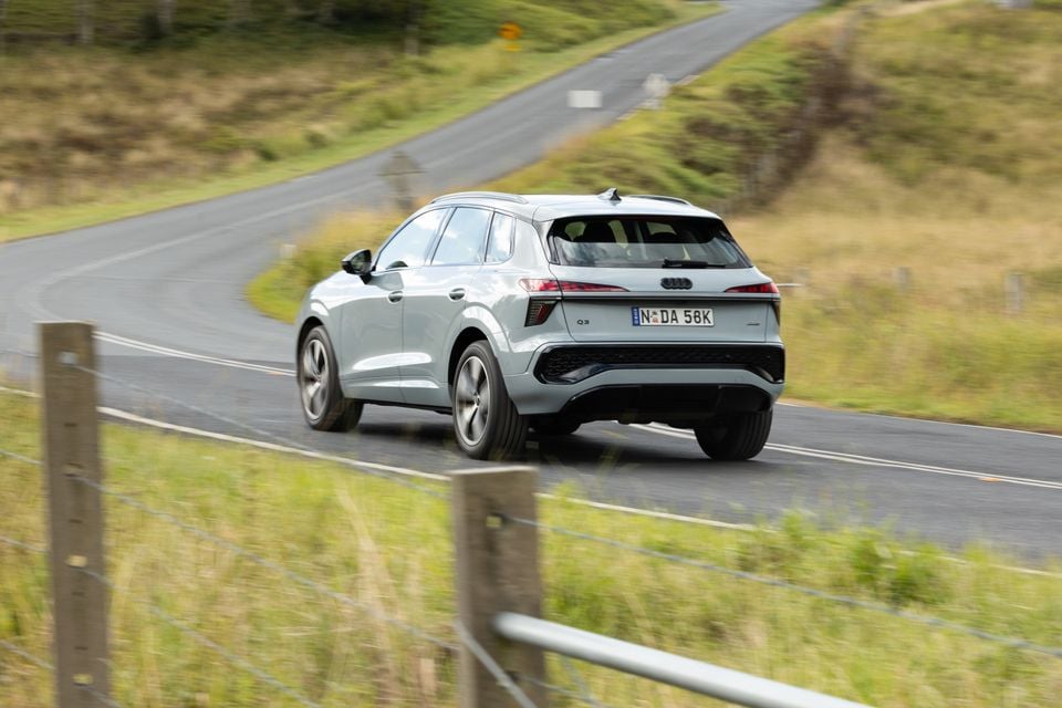 An Audi Q3 SUV in light grey driving away on a winding rural road, seen from the rear three-quarter angle