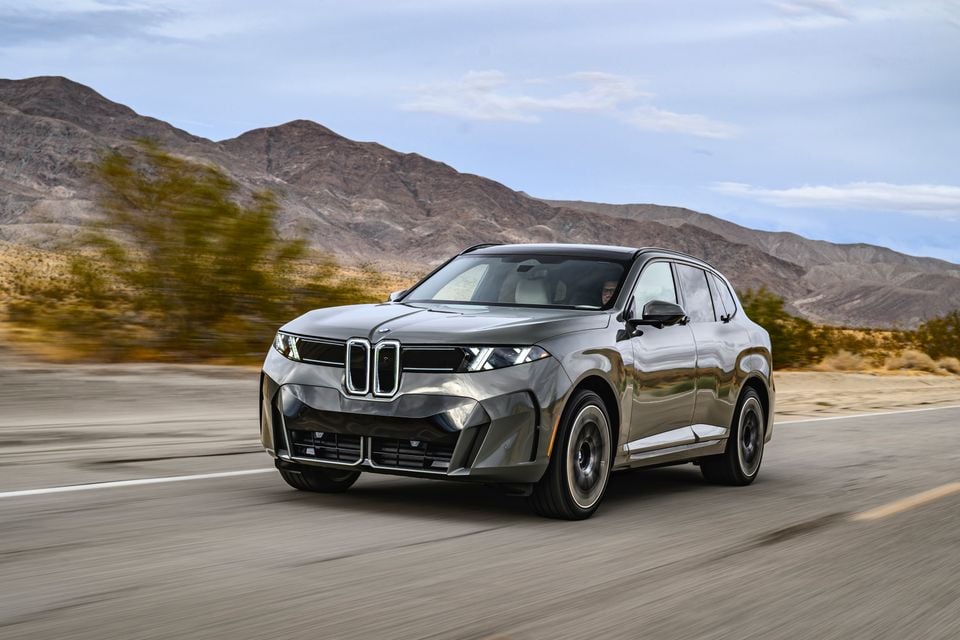 A grey BMW electric SUV driving along a desert highway with mountains behind, seen from the front quarter