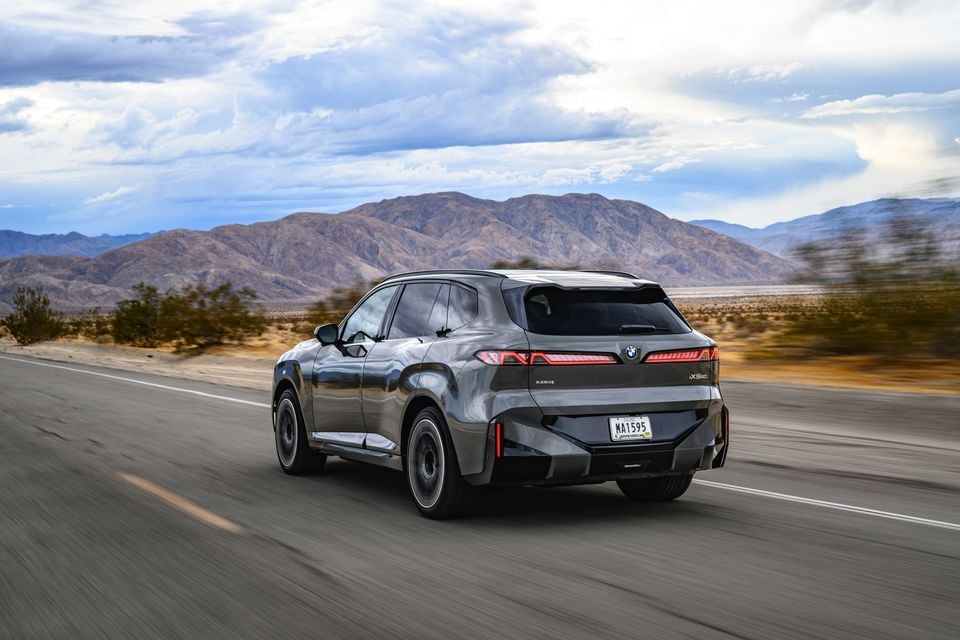 A grey BMW iX5 60 xDrive electric SUV driving along a desert highway towards mountains, seen from the rear