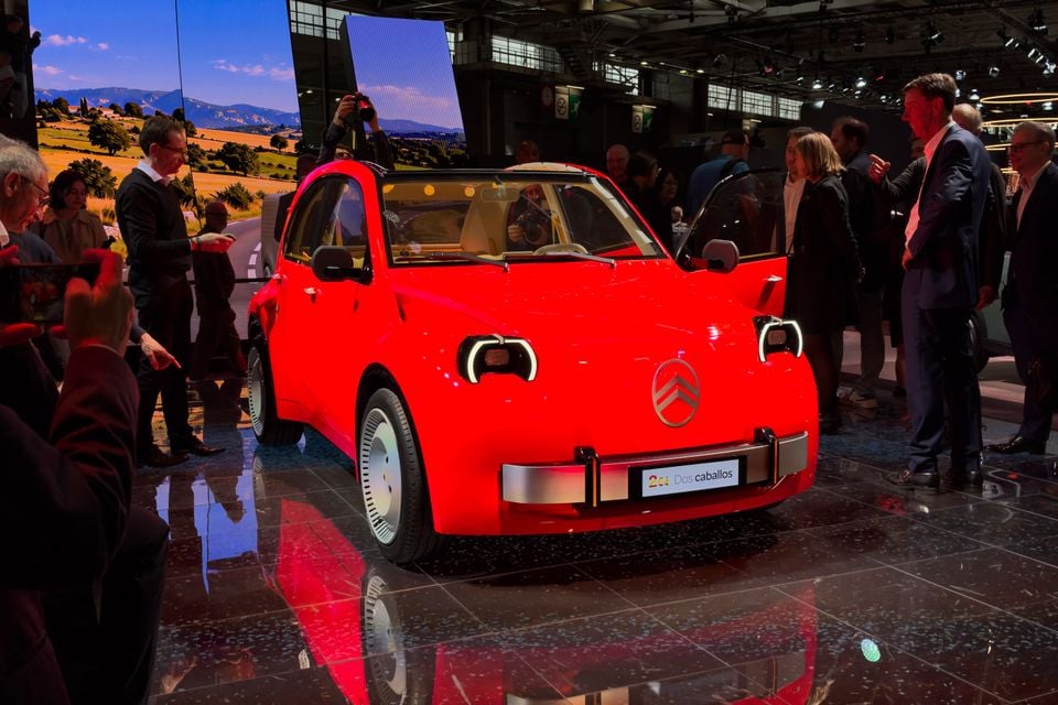 A red Citroen 2CV concept car with its door open, surrounded by onlookers and photographers on a motor show stand