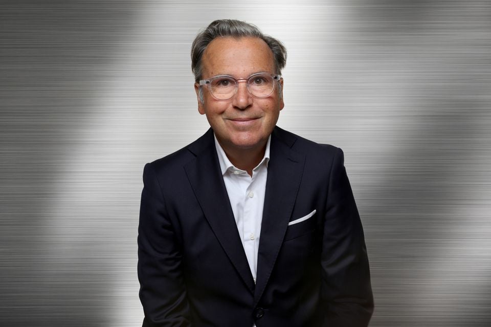Fiat CEO Arnaud Belloni smiling in a navy suit, white shirt and clear-framed glasses against a brushed metal backdrop