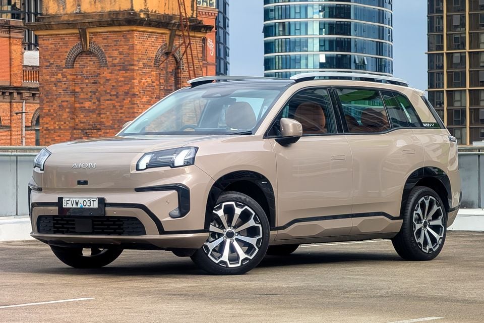 A beige GAC Aion V SUV seen from the front-side angle, parked on a rooftop car park in an urban setting with city buildings in the background.