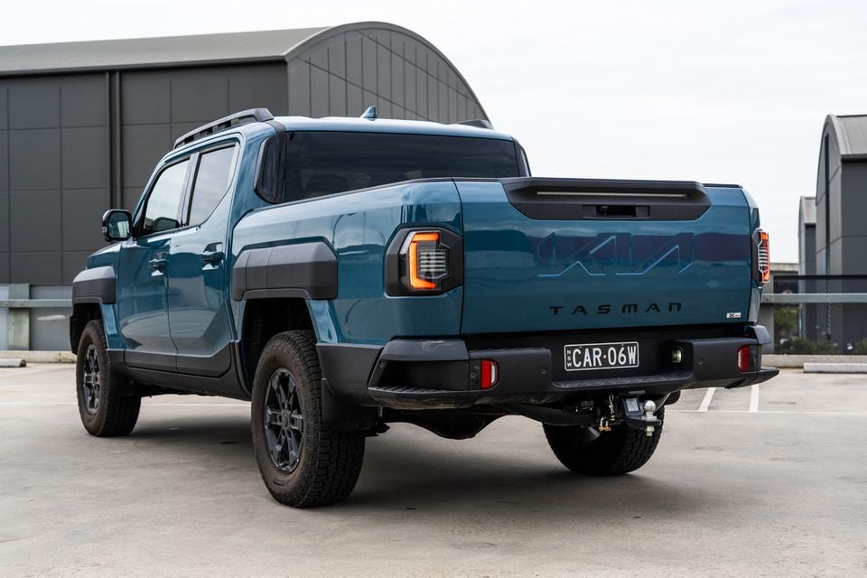 A teal Kia Tasman X-Pro dual-cab ute seen from the rear three-quarter angle in a car park, showing the tailgate badging and tow bar