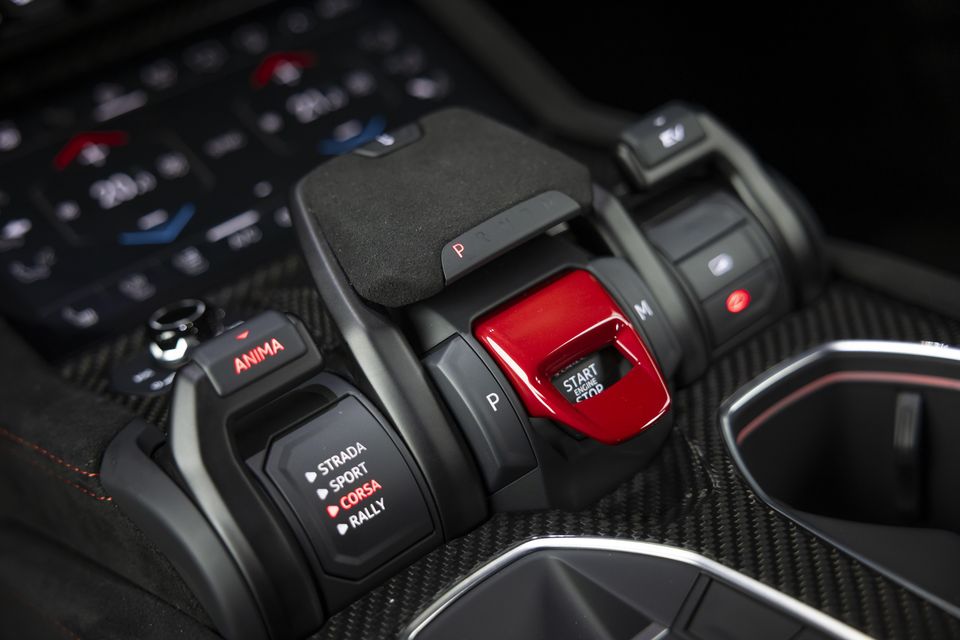Close-up of the Lamborghini Urus centre console showing the ANIMA drive mode selector with Strada, Sport, Corsa and Rally modes, a red Start Engine Stop button, and gear selector controls on a carbon fibre surround
