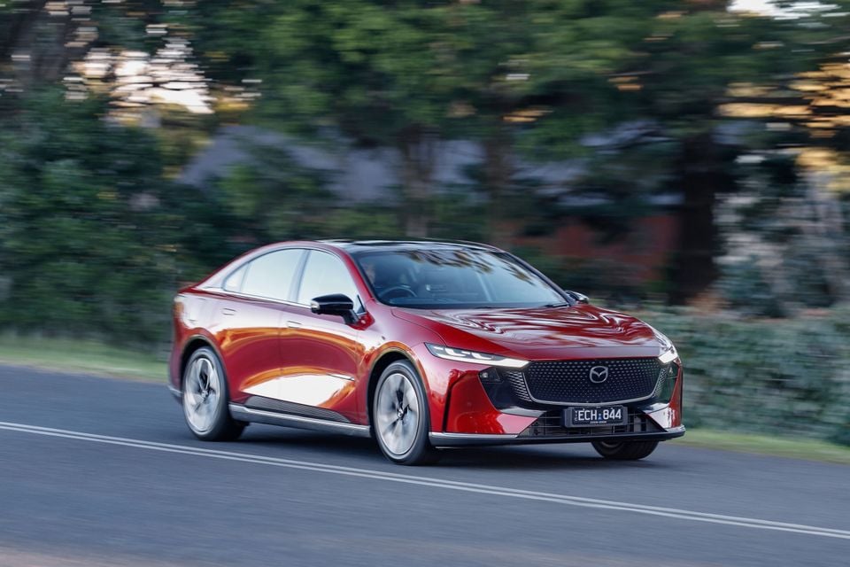 A red Mazda 6e electric sedan driving on a country road with trees blurred in the background.